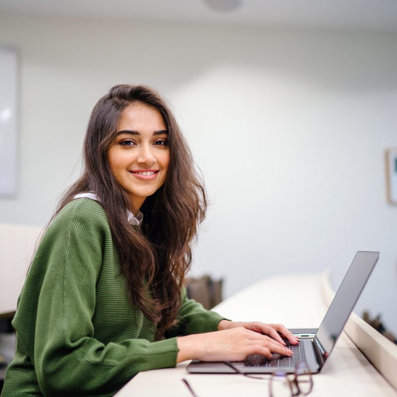 Portrait of a beautiful, young and intelligent-looking Indian Asian woman student wearing a white shirt and green tracker smiling as she works on her laptop in a university classroom.